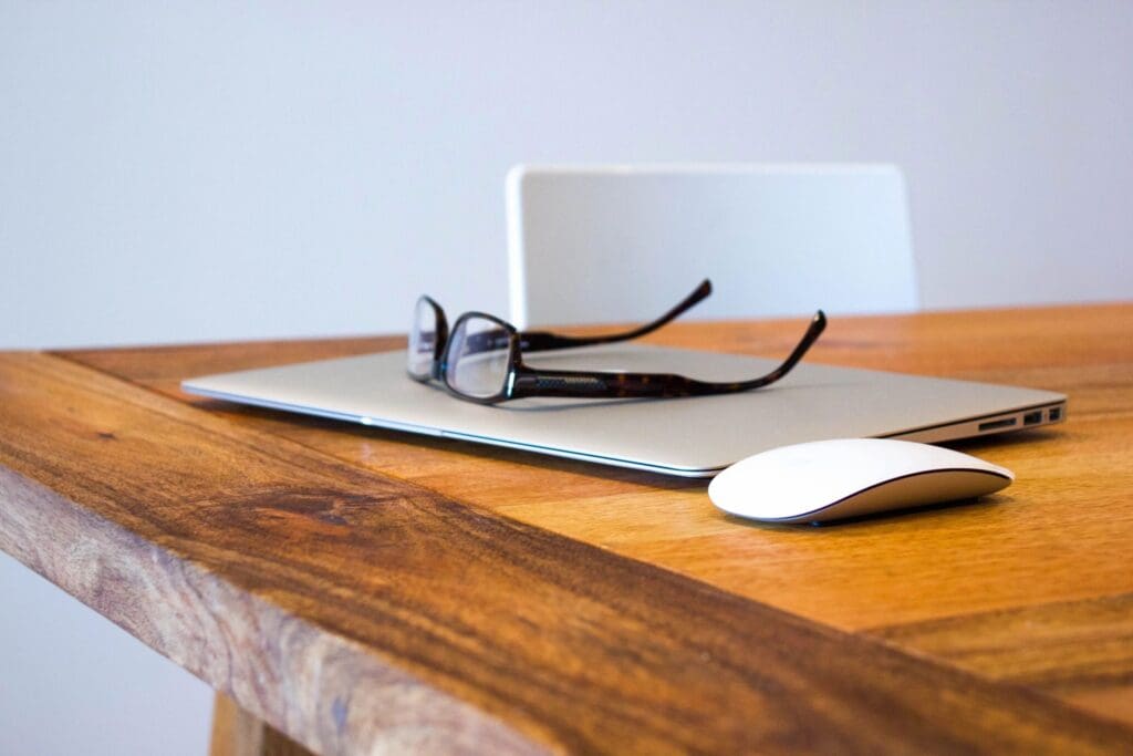 A laptop, glasses, and mouse on a wooden desk with a white chair.